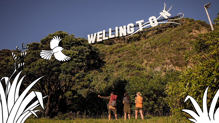 Three people standing at the base of the Miramar hill with the Windy Wellington sign, with flax and a pīwakawaka/fantail hand drawn at the corners