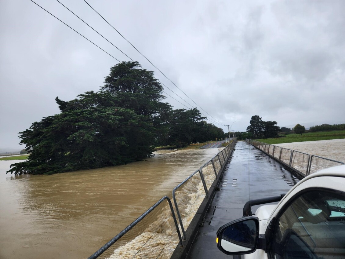 Floodwater flows alongside Jenkins Dip Bridge as viewed from a ute, with brown water close to the bridge surface and trees partially surrounded by water.