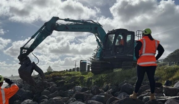 A woman in a hi-vis vest and helmet watches over a digger working on a river bank
