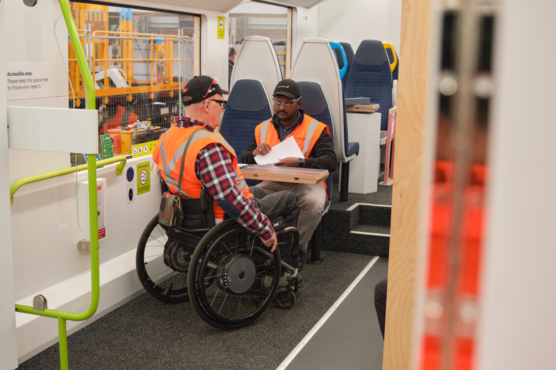Two people in hi-vis, one sat at a table and one in a wheelchair, talk inside the Tūhono mock-up train